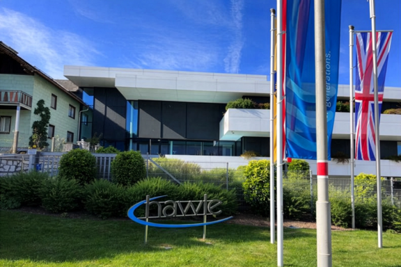 Hawle office building viewed from a landscaped lawn with a Hawle logo sign and flagpoles displaying company and national flags under a clear blue sky