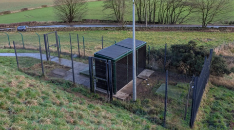 Installed LazerBooster housed within a secure fenced enclosure in a rural landscape with surrounding fields and roadside access