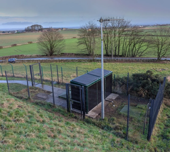 Installed LazerBooster housed within a secure fenced enclosure in a rural landscape with surrounding fields and roadside access