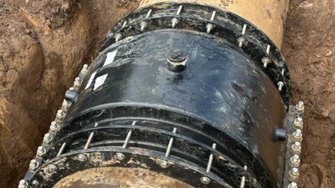 Angled view of the installed Nova Siria Duofit encapsulation collar fitted onto a large-diameter main, showing the bolted connection in the muddy ground