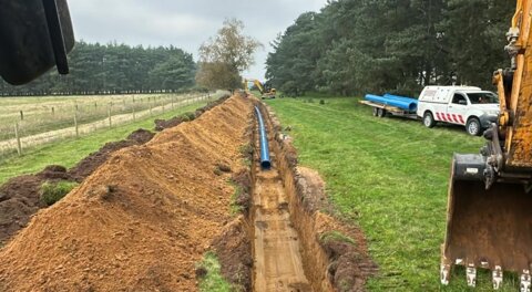 Excavated trench running through grassland, showing partially installed blue Molecor MoPVC pipeline with construction vehicles nearby