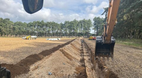 Wide view of agricultural field trench works, with excavator positioned beside a newly cut trench prepared for pipe installation