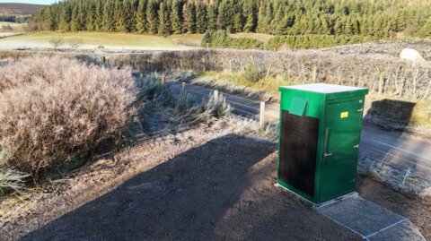 Green water quality sampling kiosk installed on a concrete base in a remote countryside setting, overlooking a narrow road with fencing