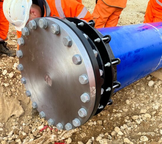 Close-up of a flange adaptor and blank flange at the end of a newly installed pipeline, with on-site workers inspecting the connection
