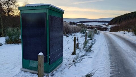 Green off-grid water quality sampling kiosk beside a snow-covered rural road, with solar panels mounted on the sides