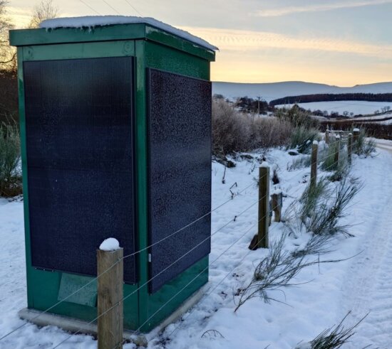 Green off-grid water quality sampling kiosk beside a snow-covered rural road, with solar panels mounted on the sides