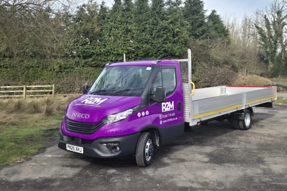 Purple R2M branded flatbed delivery vehicle parked in a rural woodland area